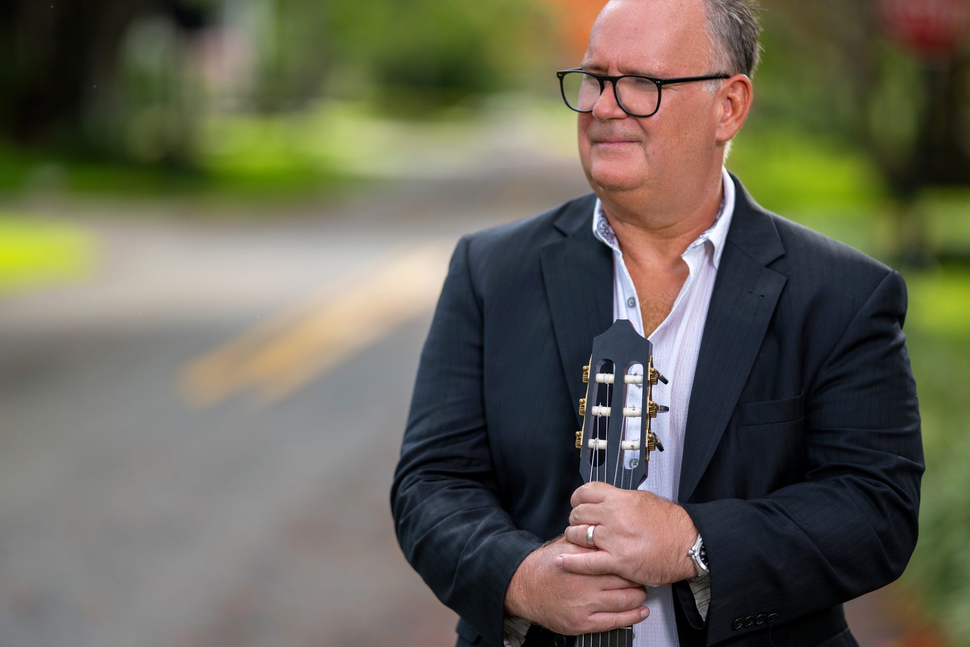 Mark Welch holding a classical guitar close with eyes closed on a tree-lined road